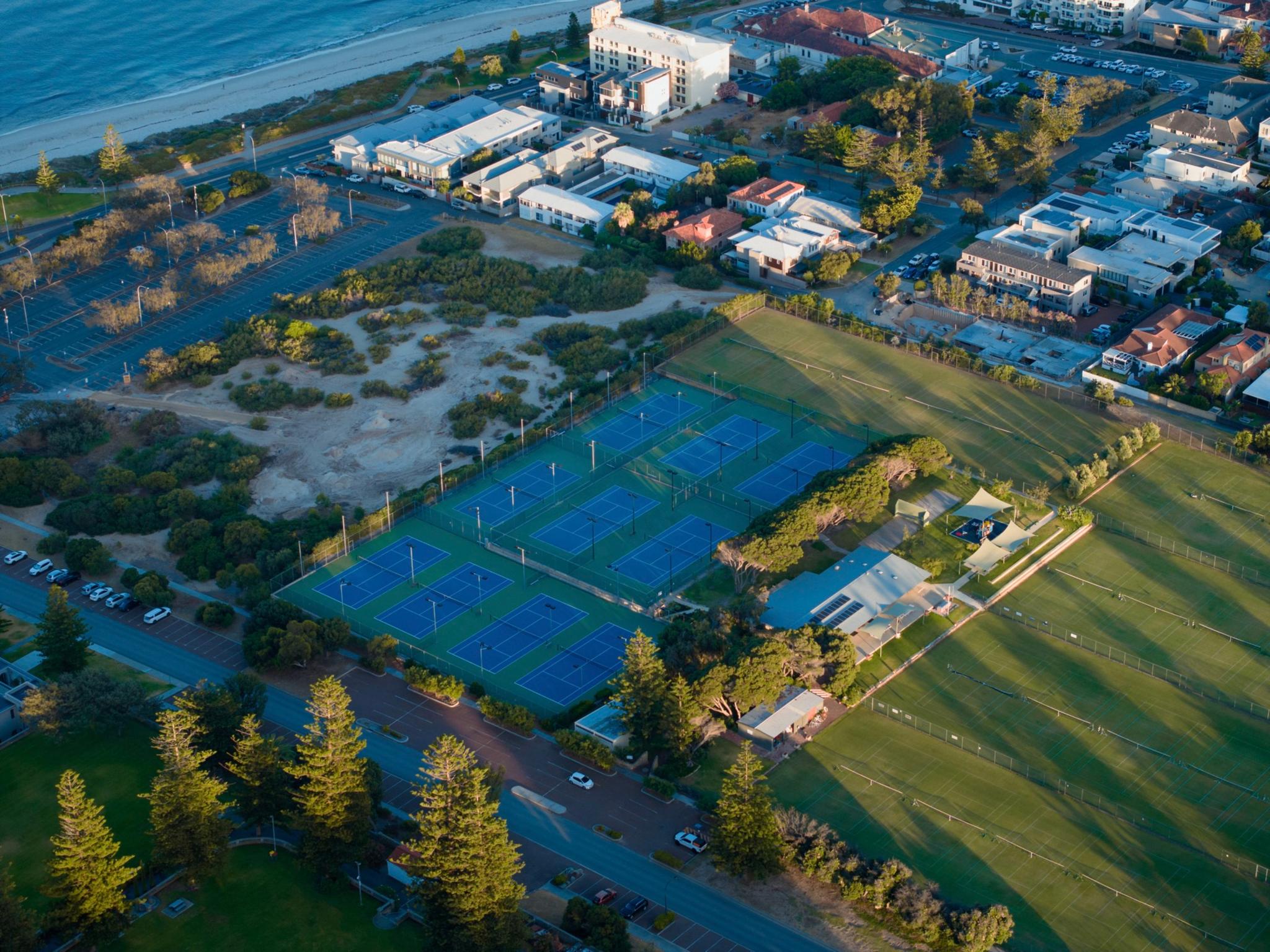 An aerial view of a coastal area featuring tennis courts, a park with green spaces, and nearby buildings The ocean is visible in the background, with a beach and a mix of residential and commercial structures Trees and pathways are scattered throughout the park area