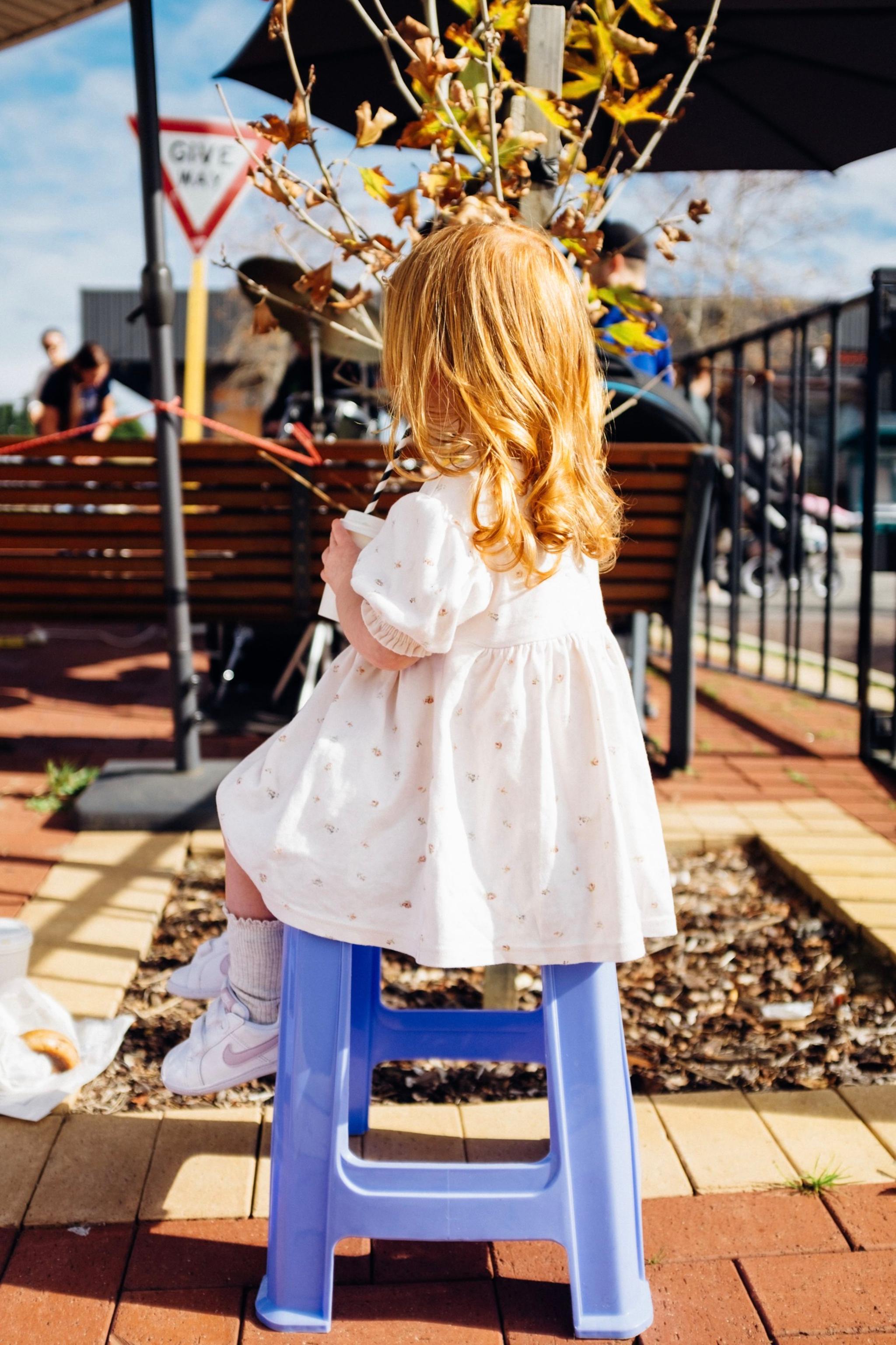 A young child with red hair, wearing a white dress, sits on a blue stool outdoors, facing away from the camera