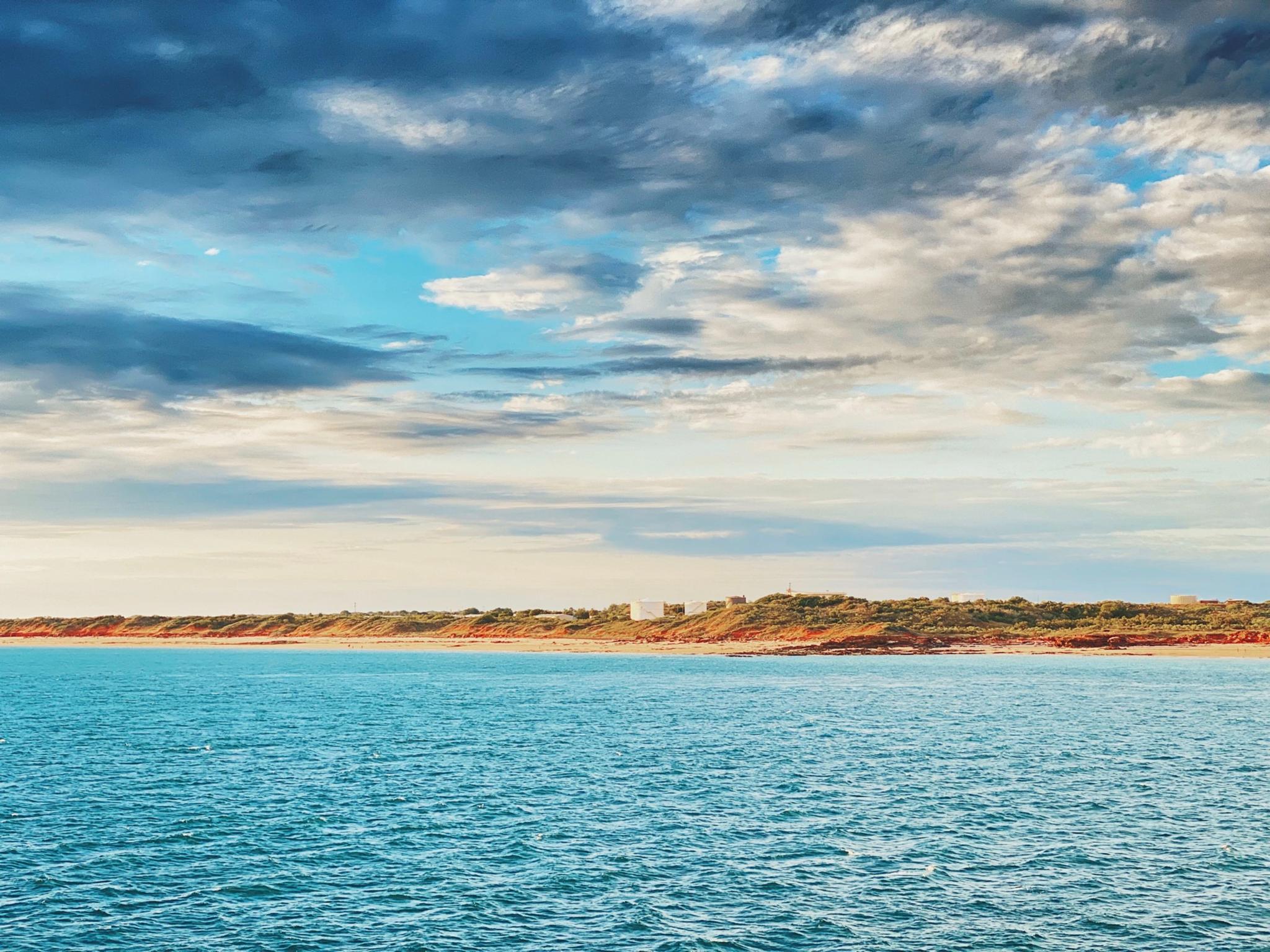 A serene coastal landscape with calm blue waters, a sandy shoreline, and a partly cloudy sky