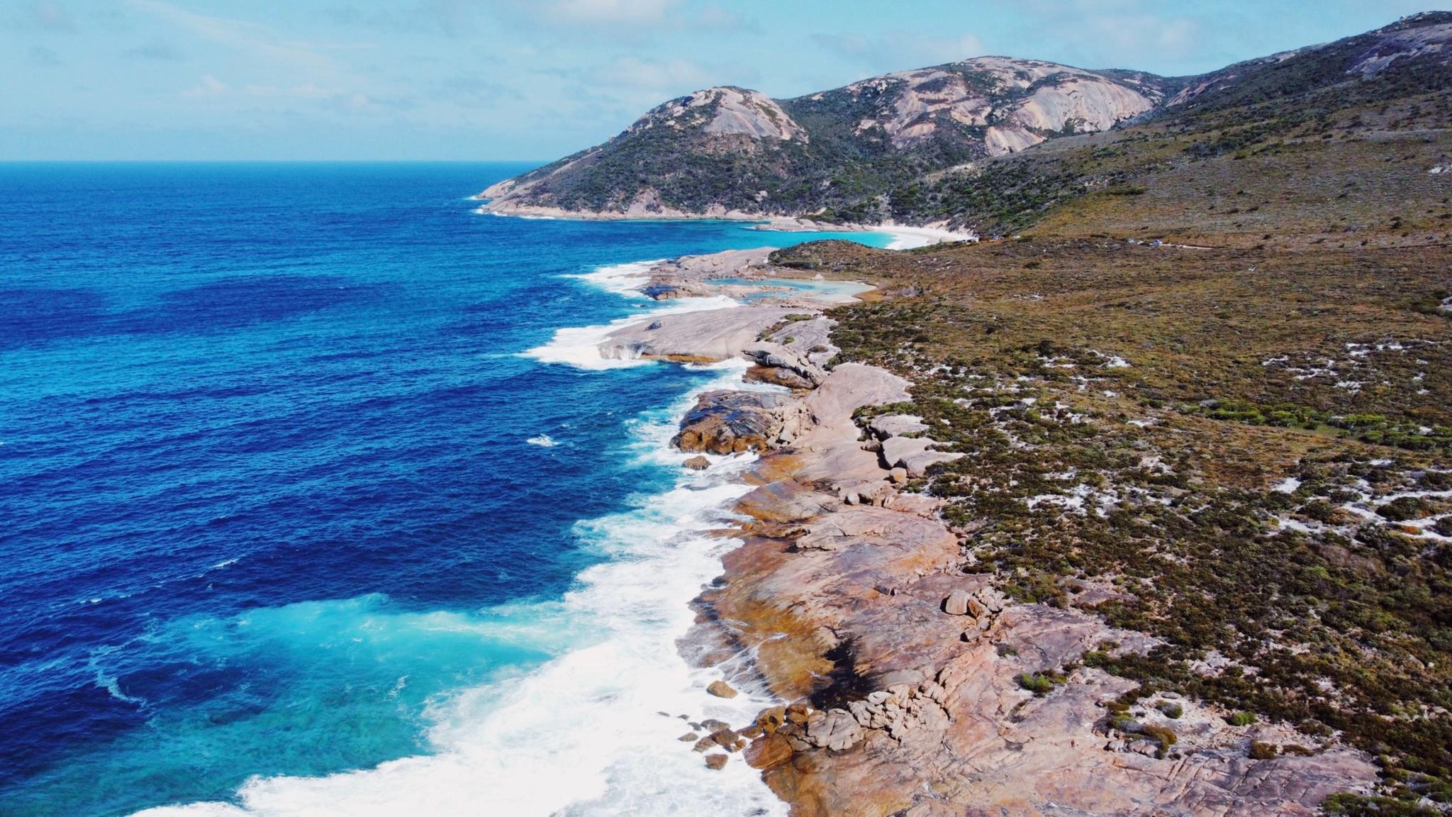 A rugged coastline with waves crashing against rocky cliffs, clear blue ocean water, and a mountainous landscape in the background