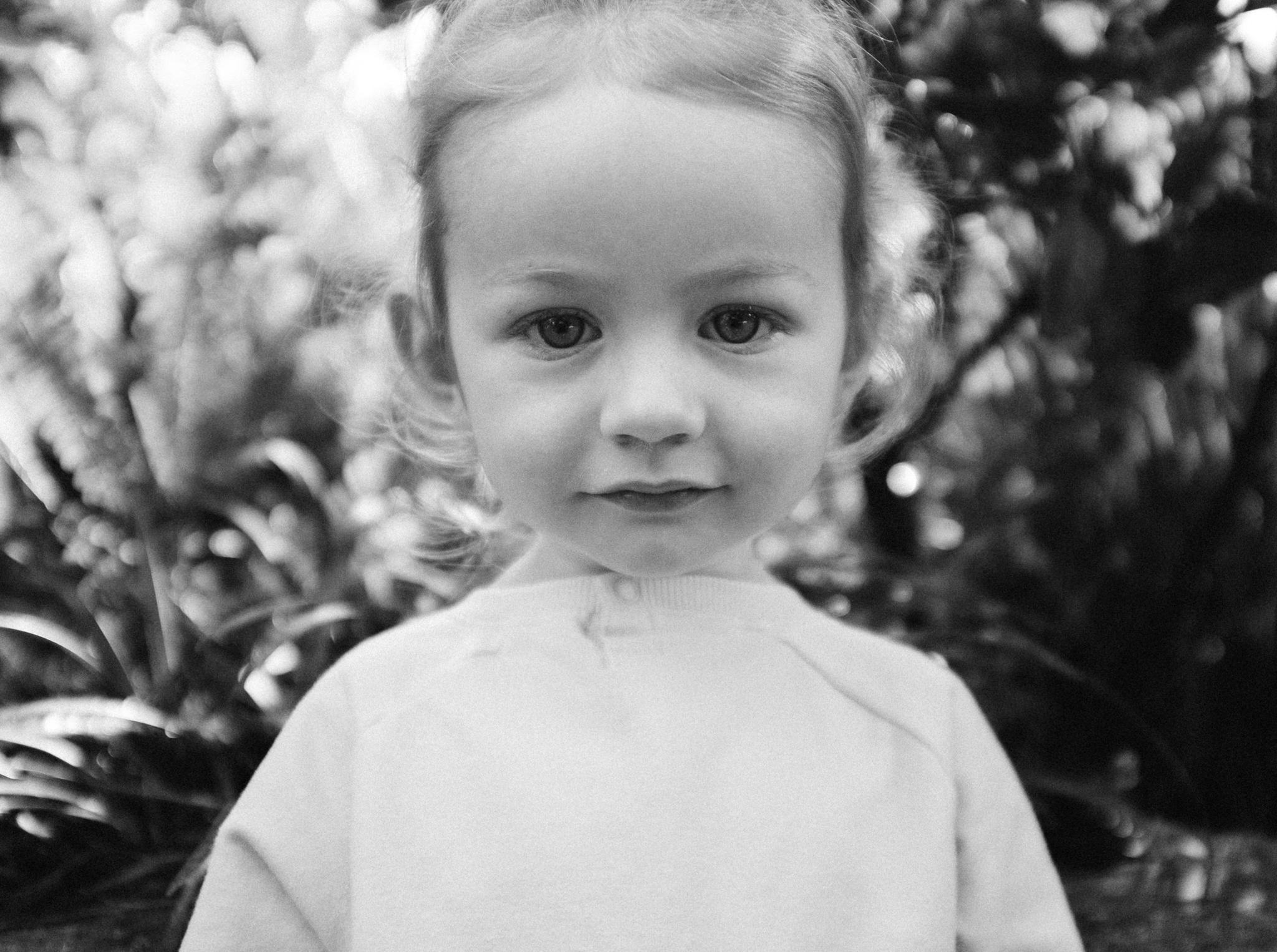 A black and white photograph of a young child with a neutral expression, standing outdoors with foliage in the background